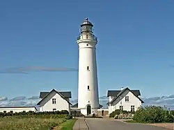 Farol entre duas construções baixas, como casas com telhado cinza típico de regiões frias onde há incidência de neve. Ambos o farol e as construções são brancos.
