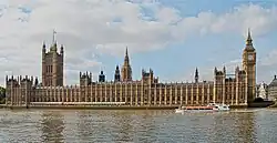 Vista do longo edifício do Parlamento, vista da margem sul do Tâmisa. À esquerda do edifício está a Torre Vitória, hasteando a Union Flag; à direita está a Torre Elizabeth (frequentemente chamada Big Ben).