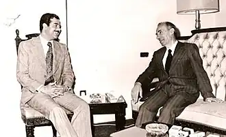 Saddam Hussein and Michel Aflaq seated, talking. Both wear suits.