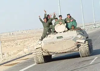 Six uniformed soldiers waving from an armoured vehicle on a highway