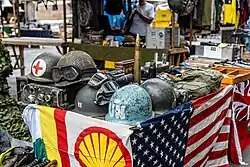 Mesa na Feira de Antiguidades da Praça XV. Há artigos militares históricos, incluindo capacetes militares (um com cruz vermelha de médico, outro com rede camuflada e um azul-claro da ONU), equipamento de rádio antigo, óculos de proteção e uma munição de artilharia. Na frente da mesa, há bandeiras decorativas, como a bandeira da Shell e a bandeira americana, além de parte de uma bandeira britânica.