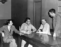 Groves sentado a uma mesa completamente limpa. Chadwick, sentado ao seu lado, observa. Tolman senta‐se do outro lado, e Smyth se inclina sobre a mesa.