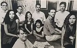 10 pessoas posam para uma foto preto e branco. A maioria está sentada ao redor de uma mesa, nela há um jornal