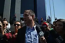 Um homem branco, trajando uma camisa azul e terno preto, é cercado por jornalistas. Uma entrevistadora do SBT põe o microfone na frente de Joesley.