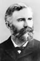 Foto de perfil em preto e branco de um homem branco com bigode e barba volumosos trajado formalmente. O mesmo está olhando para esquerda com uma expressão facial que não demonstra emoção.