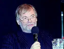 Gaardner é um homem branco de cabelos e barba grisalhos e usa óculos de grau. Aqui ele fala em um microfone.