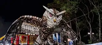 A imagem mostra um carro alegórico da agremiação GRCES Unidos de Jucutuquara, é possível ver a frente do carro, uma grande escultura de uma coruja, símbolo da escola, vestida em uma armadura medieval oriental, no fundo, uma construção no estilo oriental com várias lanternas orientais ao redor, simulando a imagem de um pequeno mosteiro.