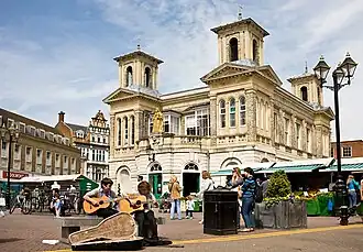 imagem de um edifício de dois andares com torres e varanda. Barracas de mercado na frente e, em primeiro plano, dois artistas de rua tocando violão.