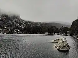 El agua contiene decenas de ondas provocadas por el viento. Al fondo de la foto se ve la cima de la montaña tapada por la niebla, que desciende hasta casi tocar el agua. La ladera de la montaña está manchada por trozos de nieve.