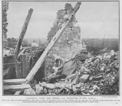 Monochrome image on newsprint type paper. Destroyed house with one remaining wall and visible roof timbers. Image of soldier dressed in British helmet and great-coat and rifle lying prone, peering over rubble towards the top right of picture.