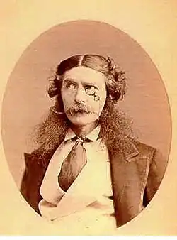 Retrato de cabeça e ombros de um cavalheiro vitoriano afetado usando um monóculo, com cabelo comprido partido no meio e encaracolado em cada lado, costeletas longas e exageradas e um bigode estilo guidão. Ele usa um casaco com lapelas muito largas, uma gola vertical e uma gravata larga enfiada em seu colete de abotoamento duplo.