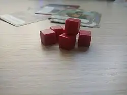Dados vermelhos empilhados em cima de uma mesa de tampo de madeira. Ao fundo, cartas do jogo espalhadas sobre a mesa.