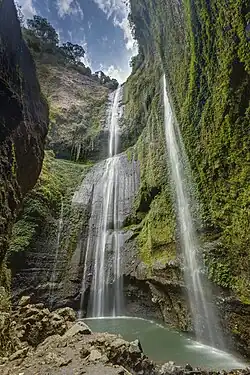 Imagem em longa exposição da Cachoeira de Madakaripura, com filetes de água caindo de um penhasco alto coberto por vegetação, criando um efeito suave e etéreo.