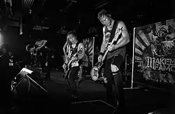 imagem em preto e branco de três músicos (homens, jovens, brancos) tocando em um palco