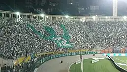 Arquibancada de um estádio de futebol lotada, com a maioria usando camisas brancas, e um mosaico em verde de um jogador apontando os dedos indicadores para o céu e com a camisa 12.