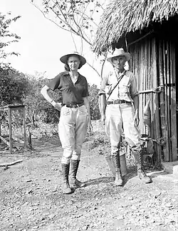 Imagem preto e branco, Matthew and Marion Stirling estão em frente a uma cabana de palha. Ambos usam calça, camisa, botas e chapéu.