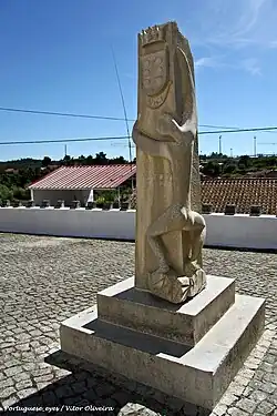Escultura vertical em calcário claro, talhada em Pedra de Ançã, com casas e céu azul ao fundo.