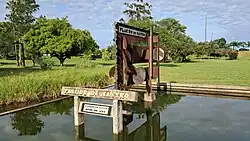 Monumento feito em aço e cimento em homenagem à Plácido de Castro, dentro de um espelho d'água. Atrás dele, há um gramado e árvores. O céu está azul com nuvens.