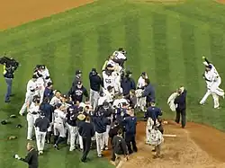 Many men in white baseball uniforms and blue caps, some wearing blue jackets, stand near a dirt mound. Some are hugging each other.