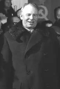 A man wearing a long coat, smiling, in front of a group of people and the Soviet flag
