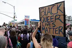 Uma placa de protesto de papelão é segurada com as palavras: "Black Queer Lives Matter" em primeiro plano, com centenas se reunindo ao fundo em um posto de gasolina Mobil