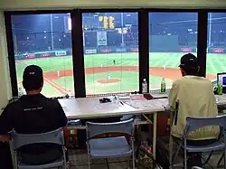 Cabine do anotador oficial, com duas pessoas observando uma partida de beisebol