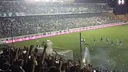 Visão da arquibancada de um jogo, onde um jogador vai cobrar um pênalti