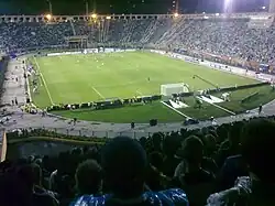 Uma partida de futebol sendo realizada em um estádio com pista de atletismo cheio, vista de trás de um dos gols; é noite e o campo está iluminado por refletores.