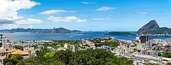 Vista panorâmica da Baía de Guanabara no Rio de Janeiro, com o Pão de Açúcar ao fundo. O céu apresenta nuvens brancas contrastando com um azul intenso.