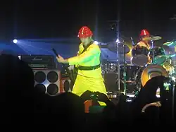 In the middle of a stage, a man wearing a yellow suit and a red hat plays a guitar. In the background is a similarly dressed man playing drums. A crowd is visible in the foreground.