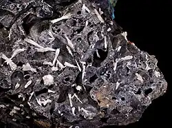 Cristais de farmacolite brancos sobre matriz escura, com estrutura fibrosa e delicada.