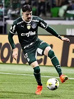 Um homem jogando futebol e com uma bola perto do seu pé esquerdo.