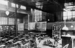 Imagem em preto e branco de uma sala ampla com diversas cadeiras dispostas em frente a uma mesa e quadros. Há grades nas janelas.