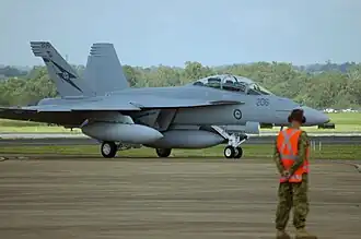 Twin-finned military jet on an airfield, with a ground crewman in an orange vest in the foreground