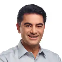 Na imagem, Raimundo Pimentel, ex-prefeito de Araripina, em uma foto de sua campanha de 2020. Ele tem cabelos pretos, curtos e bem penteados, com uma textura ligeiramente ondulada. Suas sobrancelhas são espessas e bem definidas, complementando o rosto de traços marcantes. Ele está sorrindo de maneira amigável, com dentes alinhados e expressão acolhedora. O tom de pele dele é claro, e ele veste uma camisa social de cor clara, possivelmente azul ou cinza claro, com a gola aberta e sem gravata. A iluminação é uniforme e suave, destacando os detalhes do rosto sem sombras marcantes.