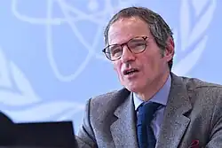 A man in glasses speaks while sitting in front of a blue background