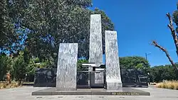 Monument consisting of two upright dark stones in front of a dark stone wall, with trees in the background