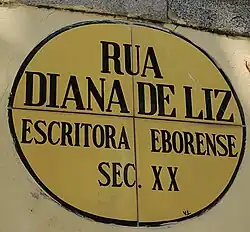 Placa oval cerâmica de fundo amarelo com linhas azuis, onde está inscrito o texto "Rua Diana de Liz. Escritora Eborense. Sec XX".