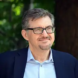 Imagem de Serhii Plokhy utilizando óculos preto, camisa azul claro e terno azul escuro com vegetação ao fundo.