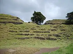 Uma série de terraços em pedra seca, parcialmente colapsados, cobertos por vegetação rasteira. No topo do terraço superior de cinco níveis permanecem os restos de dois grandes edifícios, ladeados pelas ruínas de uma estrutura menor. Uma árvore cresce à direita do edifício central menor, e outra à extrema direita, sobre o terraço superior e à frente do edifício também sobre ele