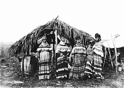Fotografia em preto e branco de quatro mulheres seminole e uma criança, de pé em frente a um chickee com padrões coloridos de algodão seminole