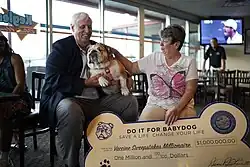 Jim Justice, a man with white hair, holding his fat English bulldog in his lap. A woman sitting to his left uses her right hand to pet the dog while holding in her left hand a large check shaped like a dog treat. The check reads "Do It For Babydog. Save A Life. Change Your Life." and has a value of one million US dollars.