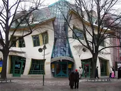 Foto da fachada da Krzywy Domek (Casa Torta), tirada em uma tarde de inverno sem neve. Um edifício de dois andares em que todas as bordas das paredes e janelas são onduladas, não há linhas retas. As duas partes de alvenaria do edifício são conectadas por um átrio de vidro com as palavras “Centrum Rezydent”. Poucos transeuntes caminham em frente ao edifício e duas pessoas ficam olhando para ele.