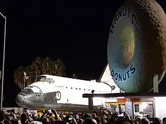 A placa da Randy's Donuts ao lado do Ônibus Espacial Endeavour enquanto ele é transportado pelas ruas de Los Angeles na sexta-feira, 12 de outubro de 2012.