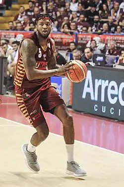 Na imagem, o jogador Julyan Stone está vestindo o uniforme do time Reyer Venezia. Ele está em ação durante um jogo de basquete, evidenciado por sua postura atlética e focada
