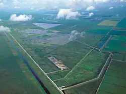 Uma fotografia aérea mostrando campos de cana-de-açúcar organizados, ladeados por canais; ao sul estão os Everglades em estado mais natural; no centro, há uma série de lagoas artificiais de cimento que atuam como áreas de tratamento de águas pluviais