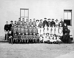 Foto em grupo posada de estudantes e professores, vestidos de preto e branco, em frente à Escola Industrial Indígena de St. Paul's, em Middlechurch, Manitoba