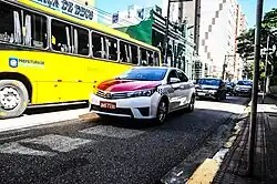 Um táxi em primeiro plano e um ônibus amarelo ao lado passam em uma rua com prédios.