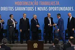 No topo da imagem, está escrito em letras maiúsculas num fundo azul "modernização trabalhista". Abaixo dela, está escrito "direitos garantidos e novas oportunidades". Encontra-se da esquerda pra direita, Ronaldo Nogueira, Eliseu Padilha, Michel Temer, Henrique Meirelles e Romero Jucá.