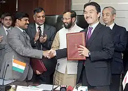 homens indianos e japoneses aplaudem, sorriem e apertam as mãos em volta de uma mesa enquanto um deles exibe uma pasta